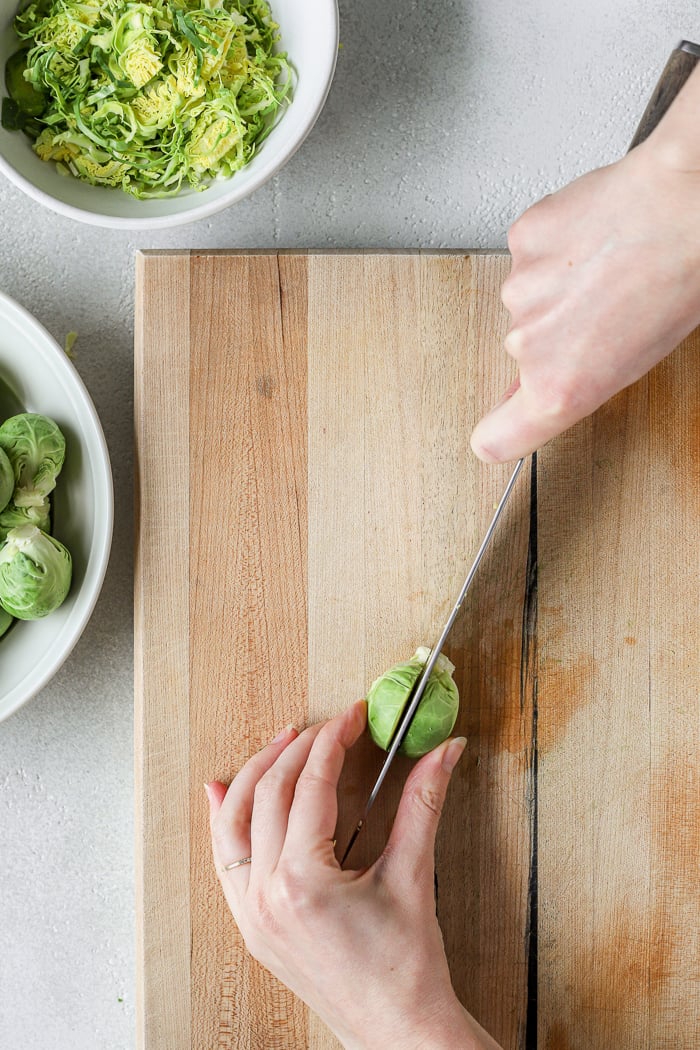 Two hands holding chef's knife cutting a Brussels sprout in half on a cutting board. Two white bowls set to the side of the image. One bowl has whole Brussels sprouts. The other bowl contains finely chopped Brussels sprouts. 