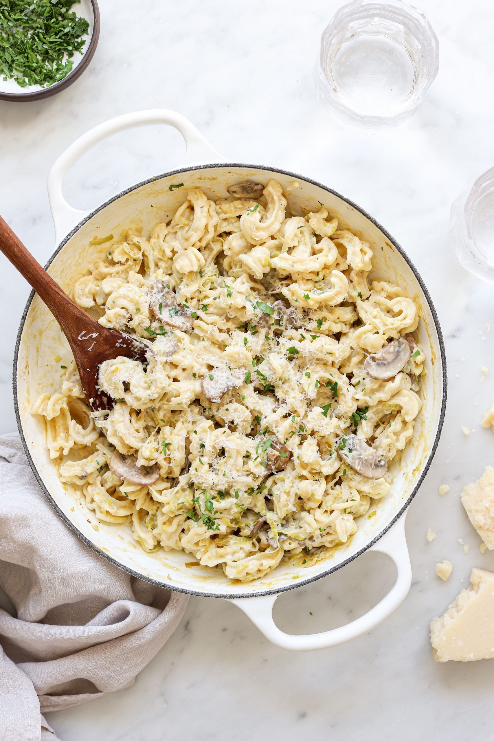 Creamy mushroom leek pasta in a large white  pasta on a table. It is garnished with more grated cheese and parsley. There is a wooden serving spoon in the pot.