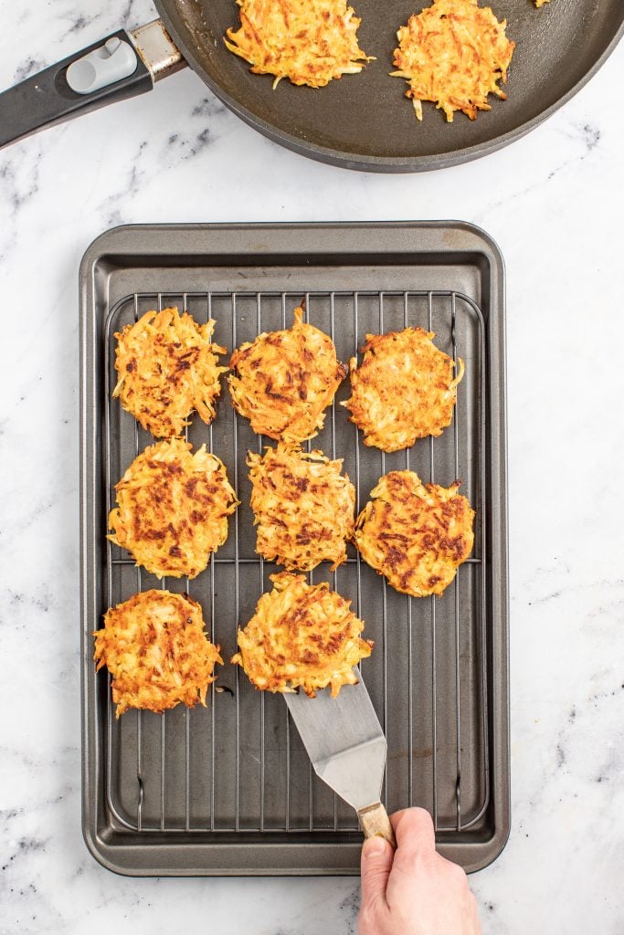 A hand using a spatula to place a cooked fritter onto a cooking rack with other cooked fritters. The cooling rack is placed on top of a baking sheet.