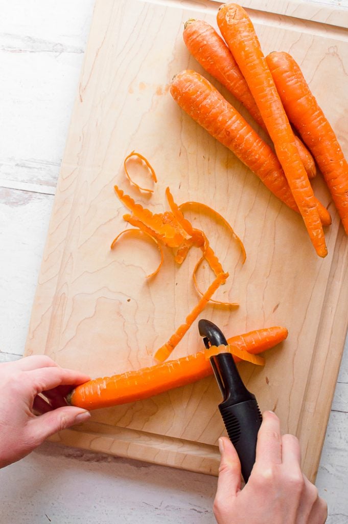 One hand holding carrot while other hand peels carrot using vegetable peeler. Vegetable peels are in the middle of a wooden cutting board. Four unpeeled carrots are set to the upper right of the cutting board. Cutting board is sitting on a white, distressed surface.