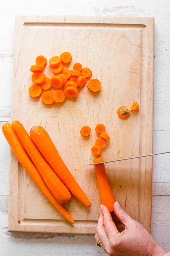 Hand holding onto a peeled carrot, being cut into coin shapes using a chef's knife. Coin-shaped carrots in a pile to the upper-left side on a wooden cutting board. Three peeled carrots are set to the left side of on a wooden cutting board. Cutting board is on a white, distressed surface.