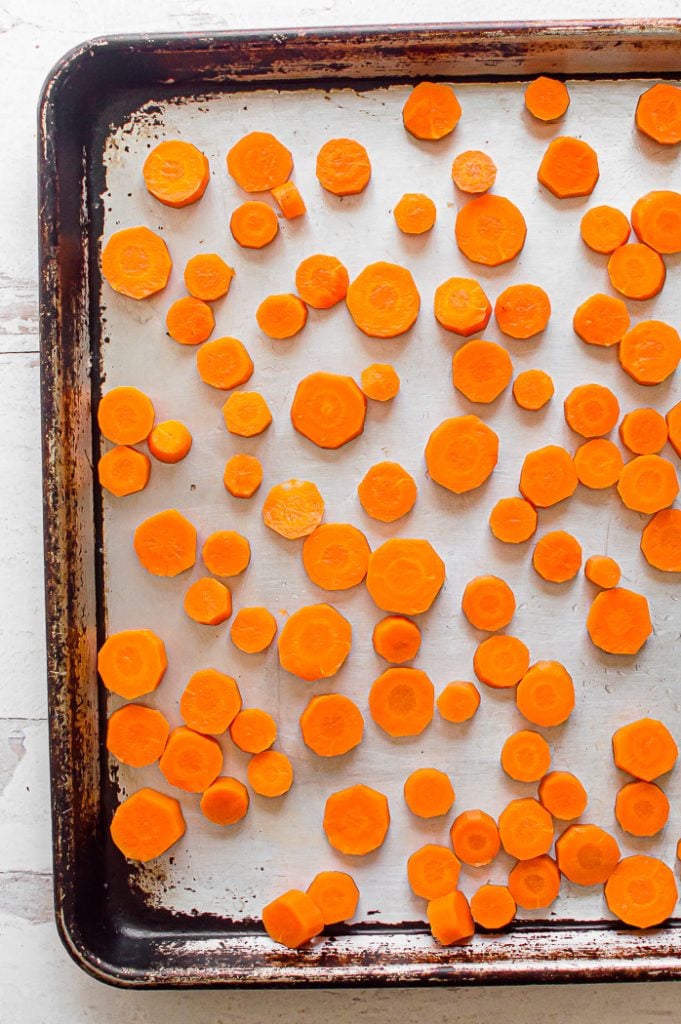 Carrots cut into coin shapes spread out on a baking sheet. Baking sheet is sitting on a distressed white surface.
