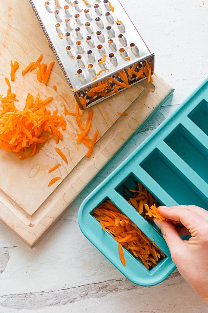 Hand placing shredded carrots into blue silicone food storage mold. Box grater with carrot remnants is sitting on top of a wooden cutting board with shredded carrots on top of the cutting board. Cutting board and food storage mold are sitting on a white, distressed surface.