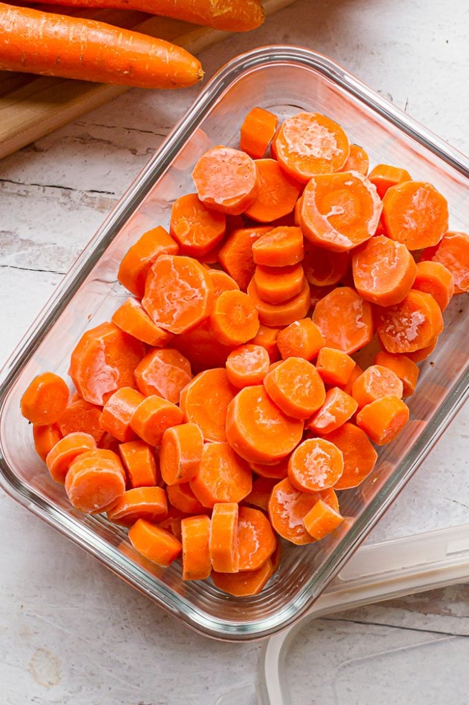 Frozen carrots in coin shape in a glass storage container on a white distressed surface. Wooden cutting board, storage container top, and carrot are off to the side mostly out of frame.