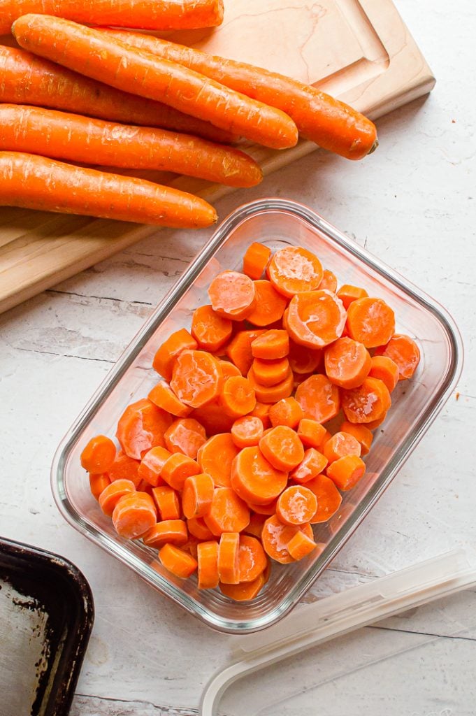 Frozen carrots in coin shape in a glass storage container on a white distressed surface. Wooden cutting board with several fresh carrots, storage container lid, and baking sheet are set aside slightly out of frame.