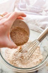 A flax egg being poured from a small bowl into a glass bowl with flour and a whisk.