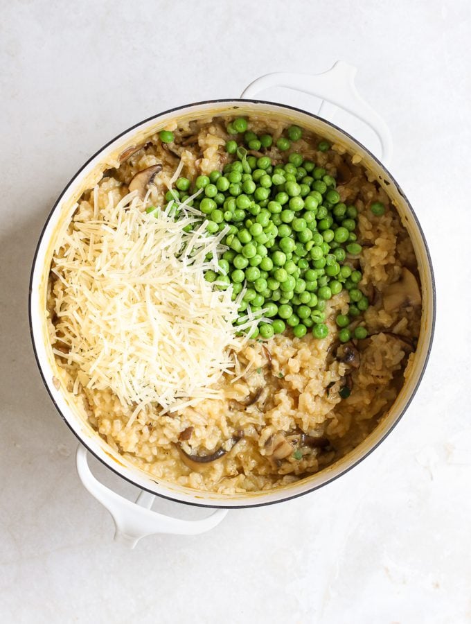 Cooked mushroom risotto in a large white pot with grated parmesan cheese and peas on top.