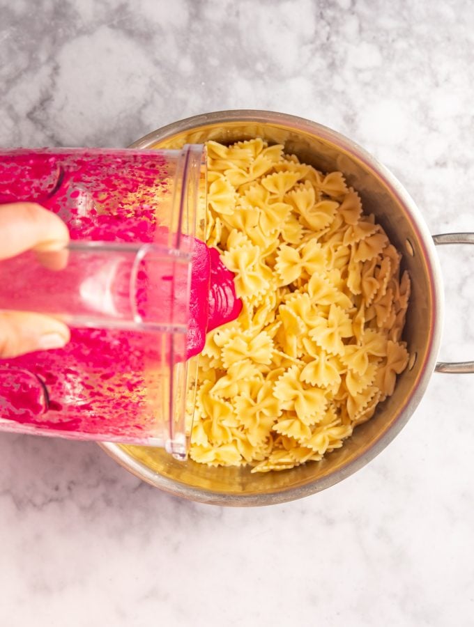 A blender with pureed creamy beet sauce being poured over a pot full of cooked bowtie pasta.
