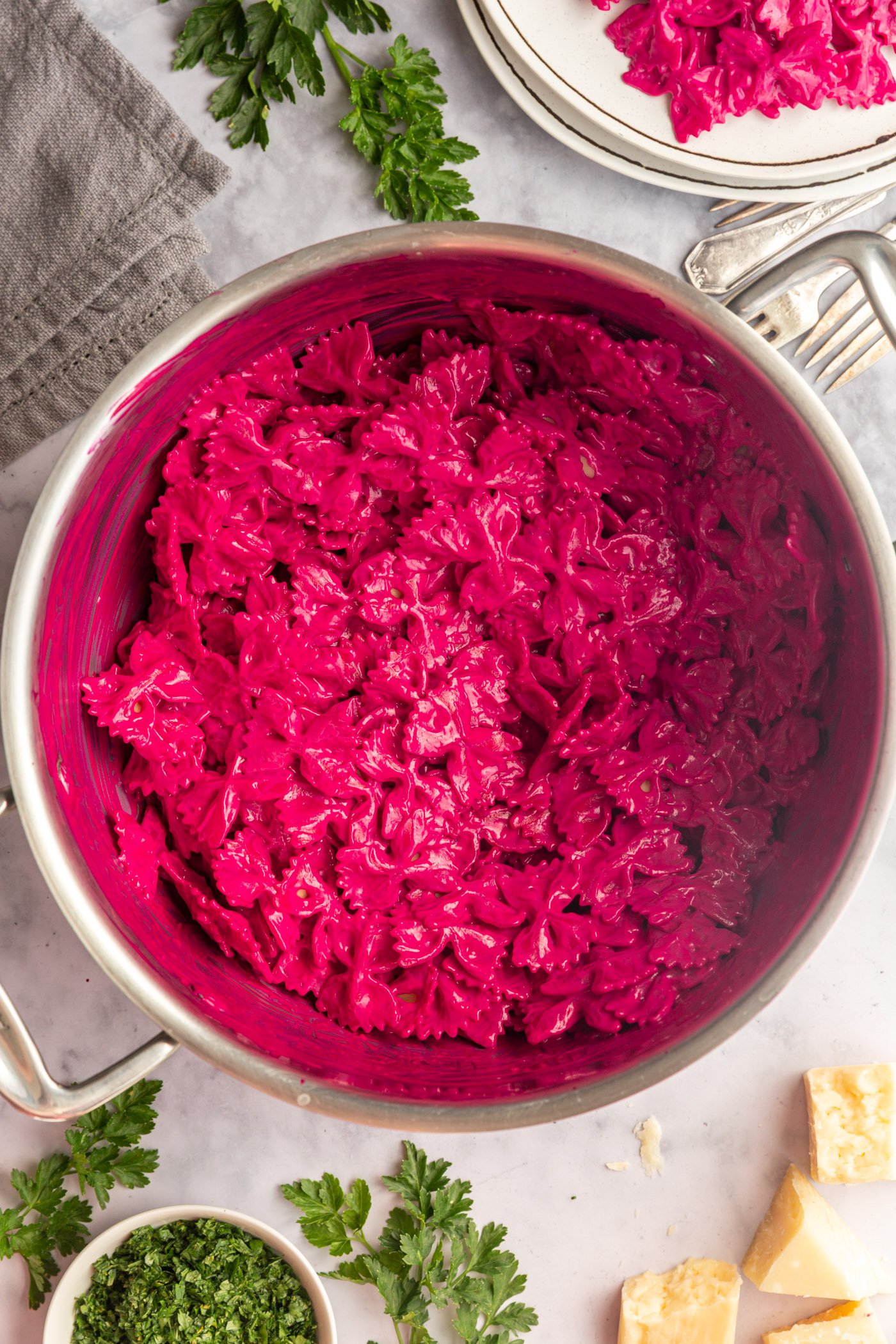 A pot of creamy beet pasta on a table with a gray napkin and parsley. There is a plate of pasta and fork off in the corner. 