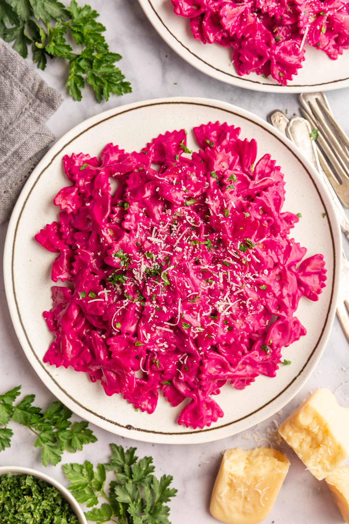 A white plate of beet pasta topped with grated parmesan cheese and finely chopped parsley. There are forks next to the plate and another plate of pasta off in the corner.