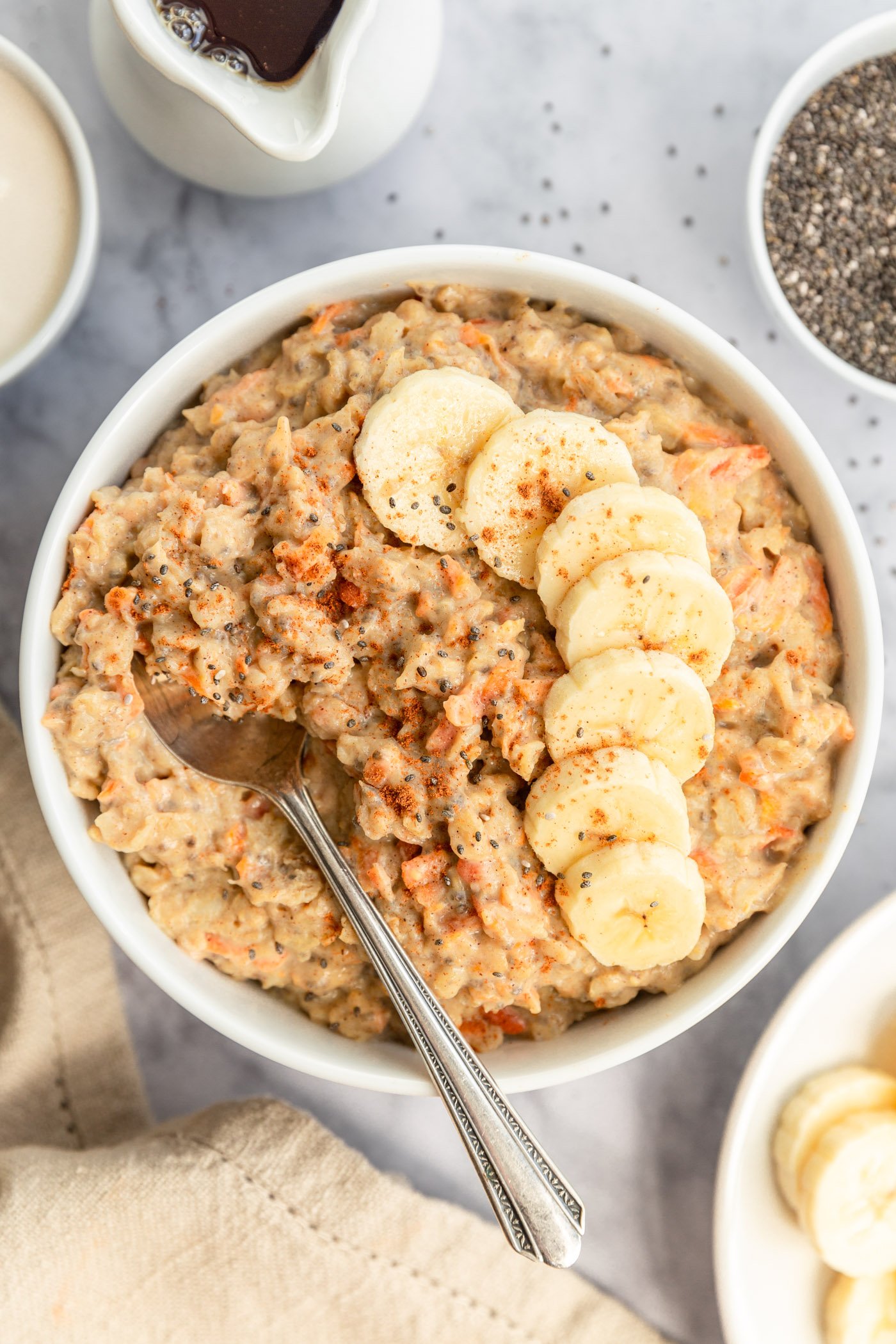 A bowl of carrot banana protein oatmeal in a white bowl on a table. There are sliced bananas and chia seeds on the top of the oats and there is a spoon in the bowl. There is a napkin and bowls of ingredients next to the bowl. 