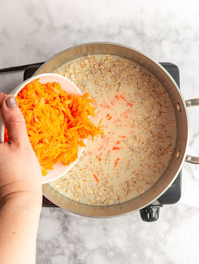 A hand pouring grated carrots from a white bowl into a pot of cooking oatmeal.