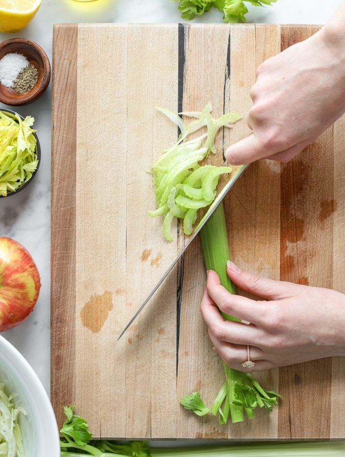 A person cutting a stalk of celery at an angle on a wooden cutting board with a knife.