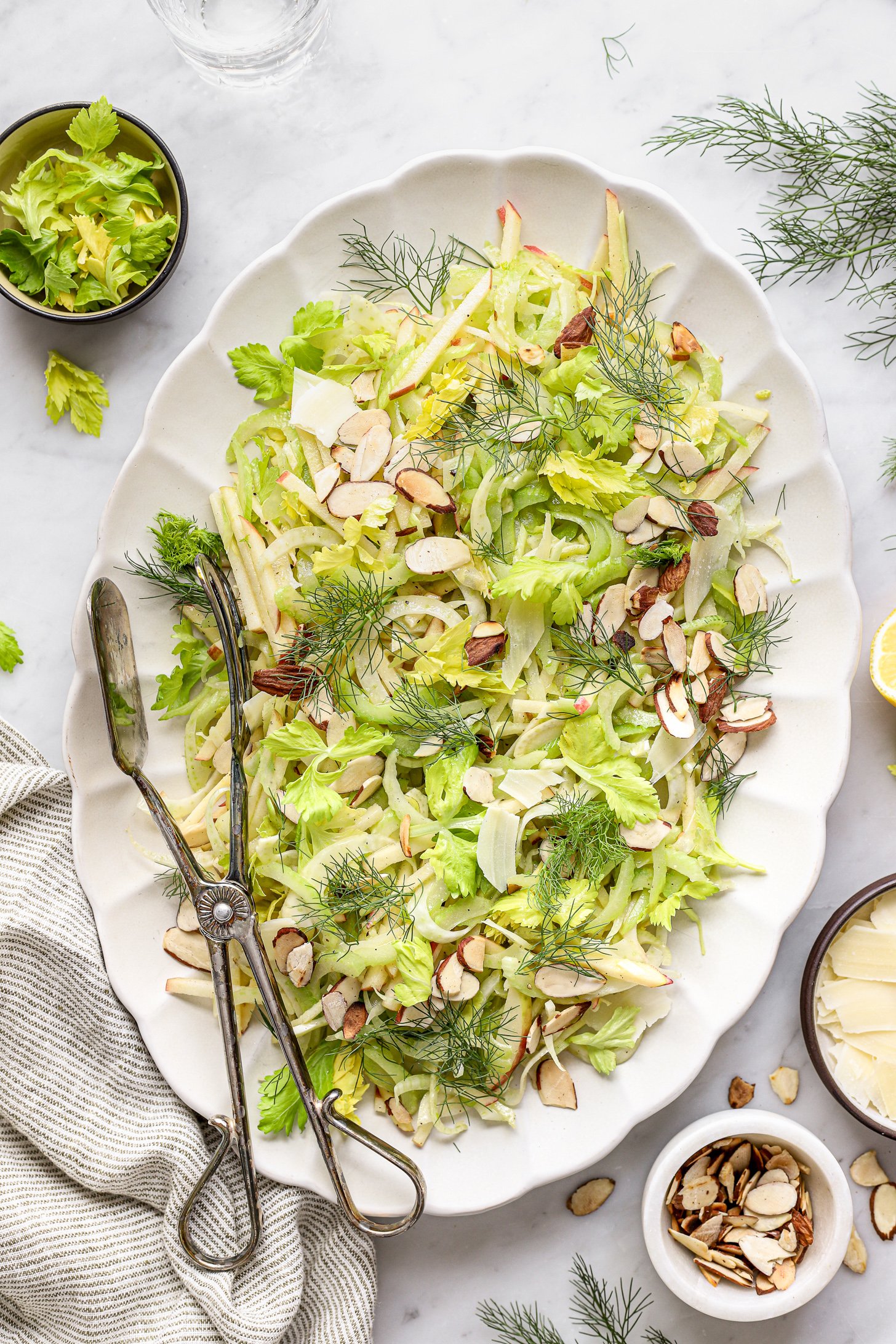 Fennel Apple & Celery Salad on a white platter with silver tongs.  There is a striped napkin and bowls of ingredients on the table next to the platter. 
