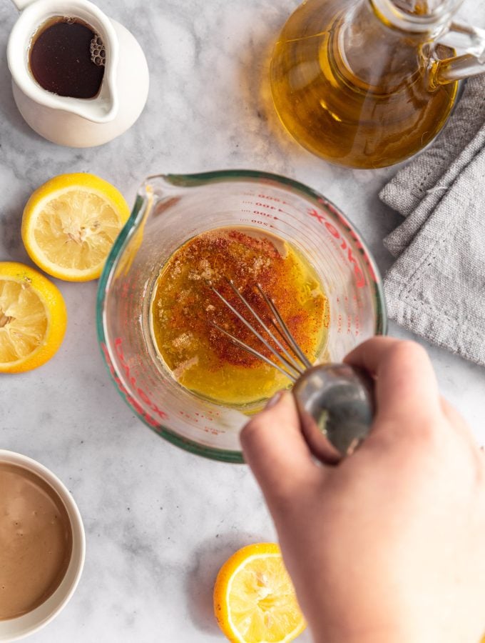A hand whisking together the ingredients for maple tahini dressing in a large glass measuring cup. There are sliced lemons, pitchers of maple syrup and oil, and a bowl of tahini next to the measuring cup.