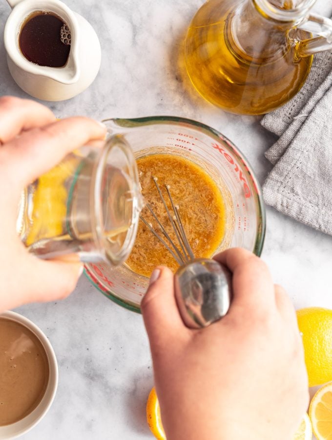 A person whisking together the ingredients for maple tahini dressing in a large glass measuring cup while also pouring water into the measuring cup. There are sliced lemons, pitchers of maple syrup and oil, and a bowl of tahini next to the measuring cup.