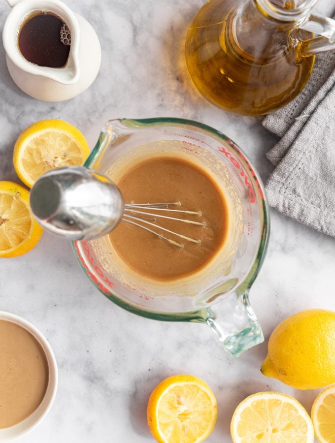 A whisk in a large glass measuring cup containing maple tahini dressing. There are sliced lemons, pitchers of maple syrup and oil, and a bowl of tahini next to the measuring cup.