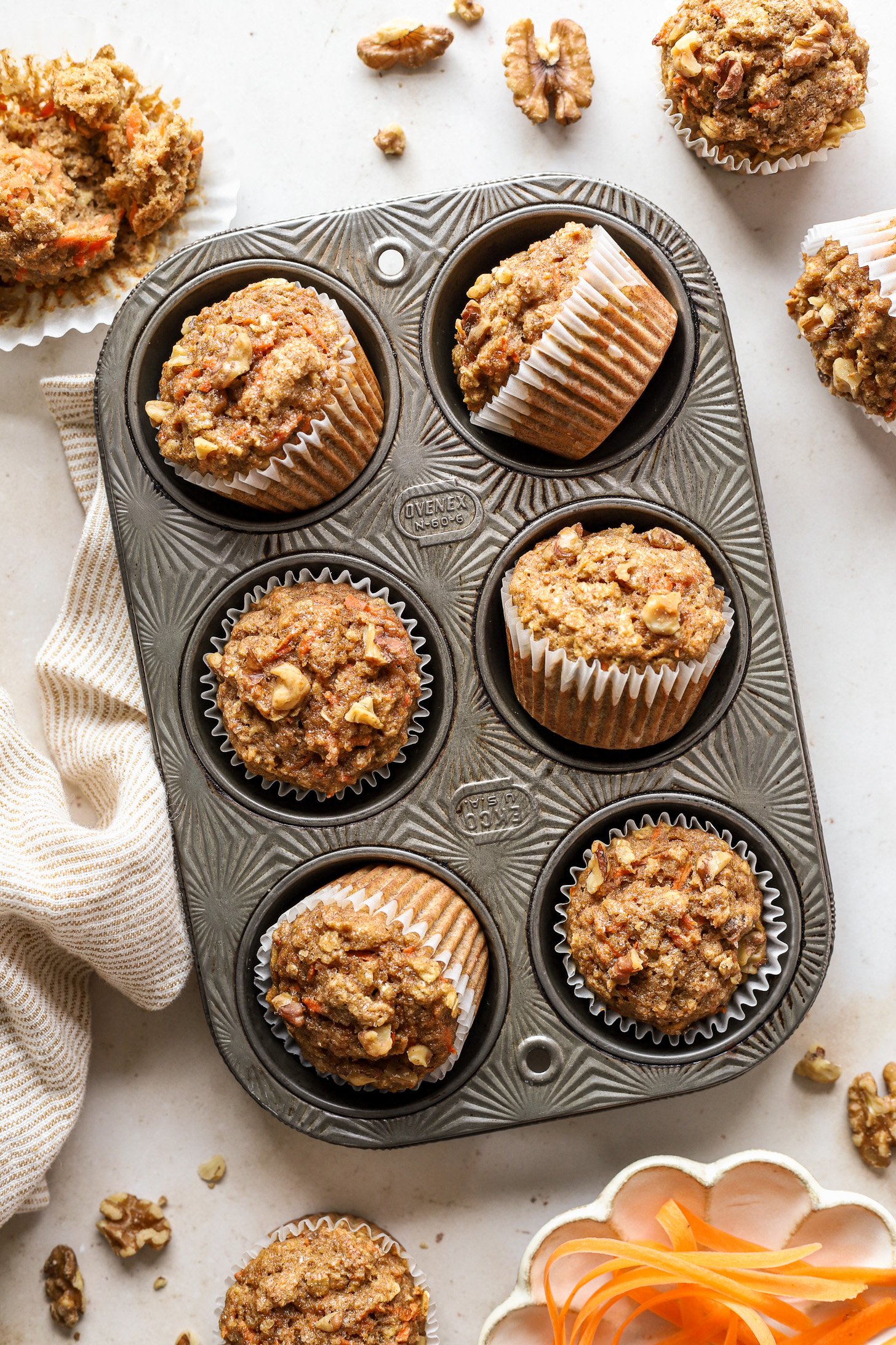 Healthy oatmeal carrot muffins in a muffin tin after baking. There are a few muffins on the counter around the tin and a striped napkin until the muffin tin.
