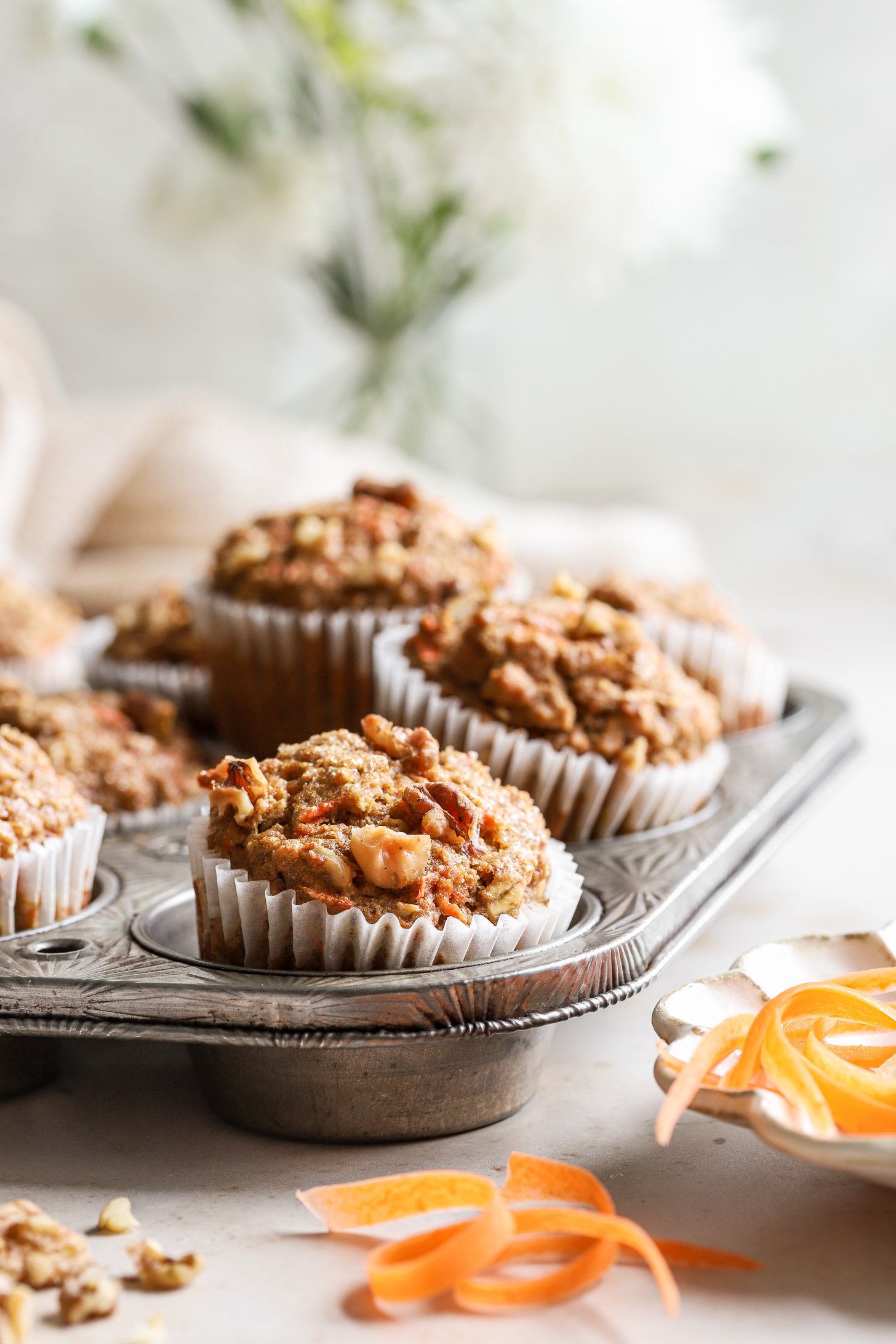 Healthy carrot oatmeal muffins in a muffin tine after baking. There are carrot peelings and walnuts scattered around the table near the tin. 