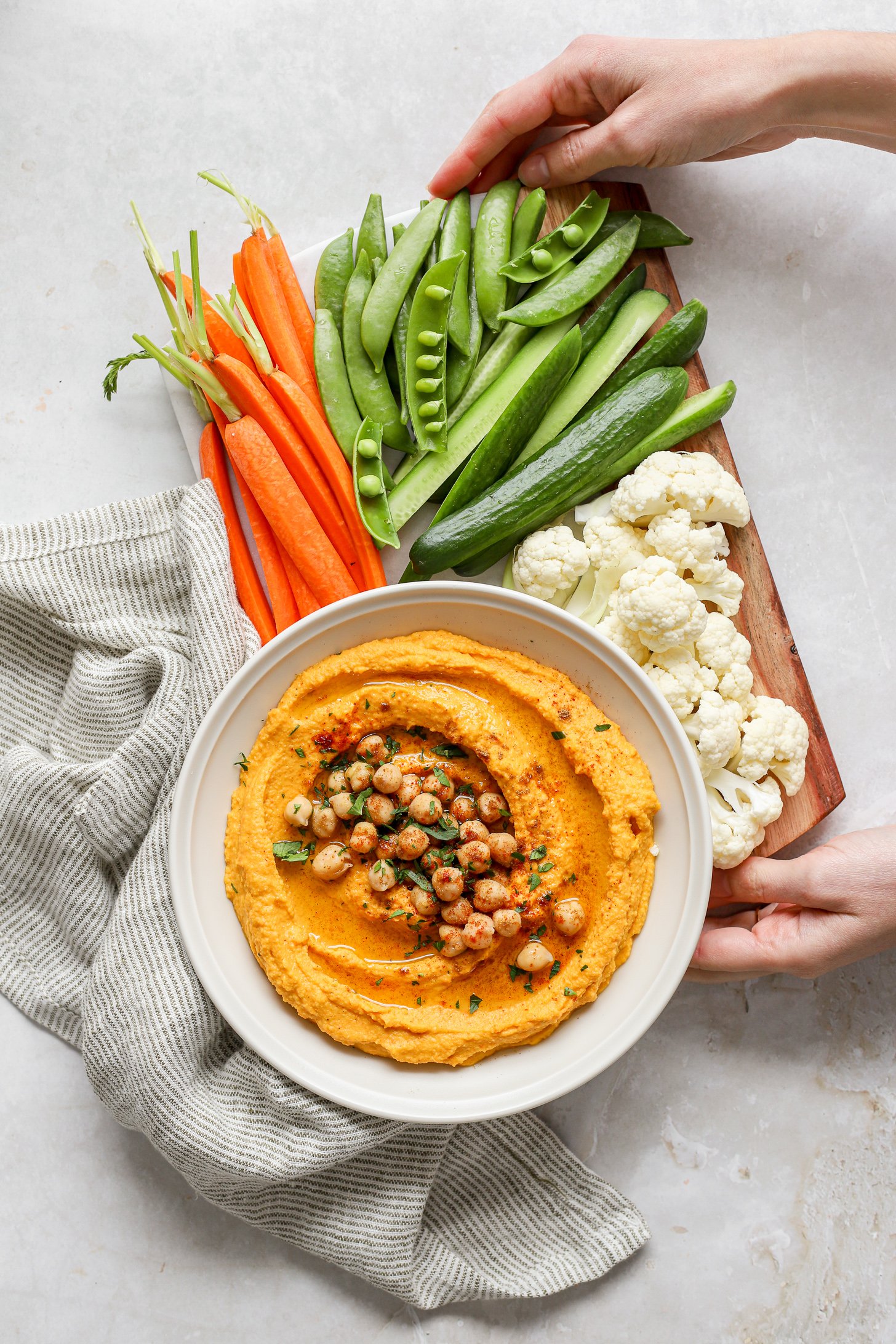 Roasted carrot hummus on a veggie board with carrot, snap peas, sliced cucumbers, and cauliflower florets. The hummus is topped with whole chickpeas and fresh herbs. There is a striped napkin next to the board and two hands holding the board.  