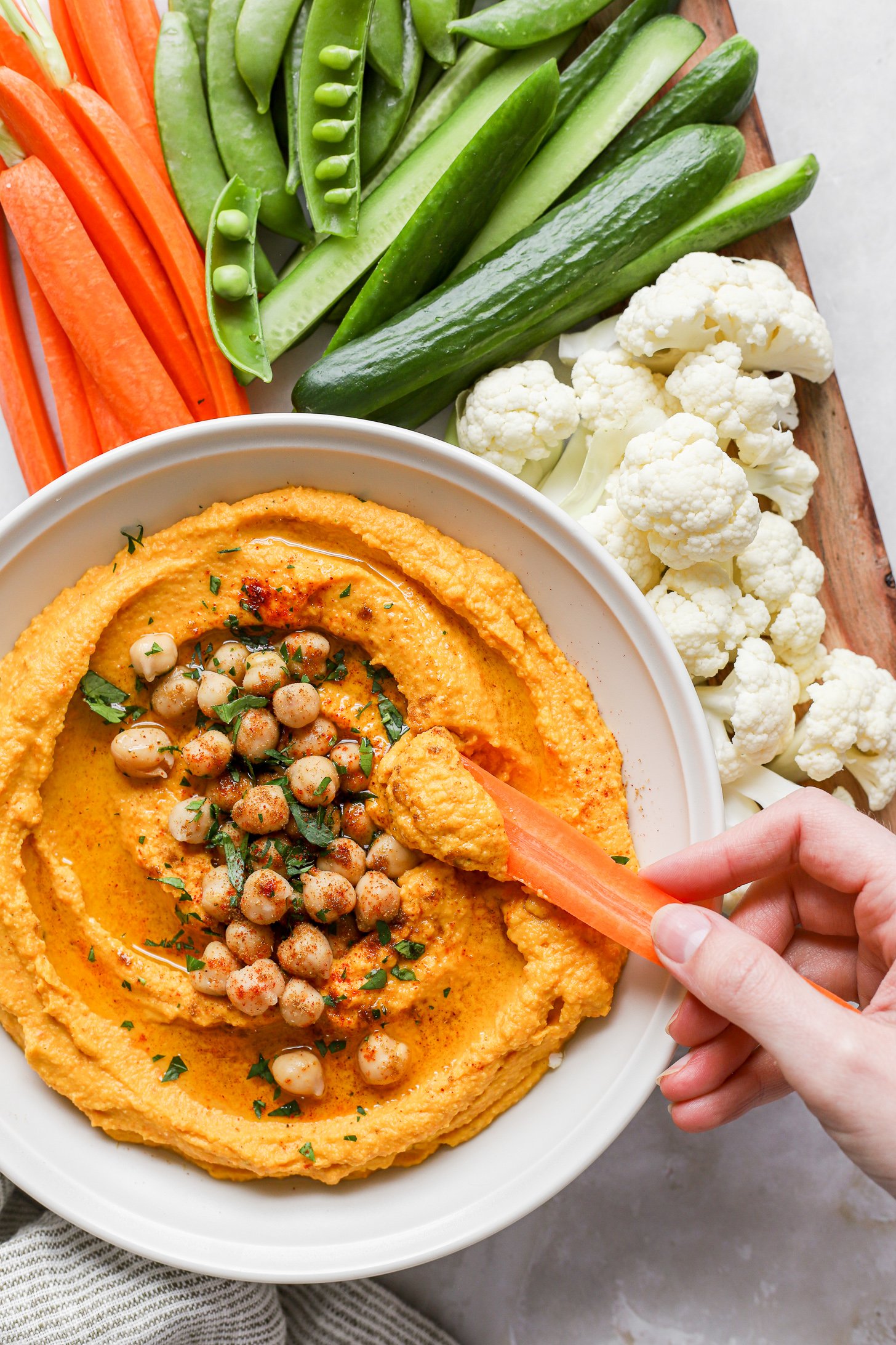 Roasted carrot in a white bowl topped with chickpeas and fresh herbs. A hand is dipping a carrot stick in the hummus and the bowl is surrounded by other sliced veggies, like cauliflower florets, sliced cucumbers, sugar snap peas and more carrot sticks.