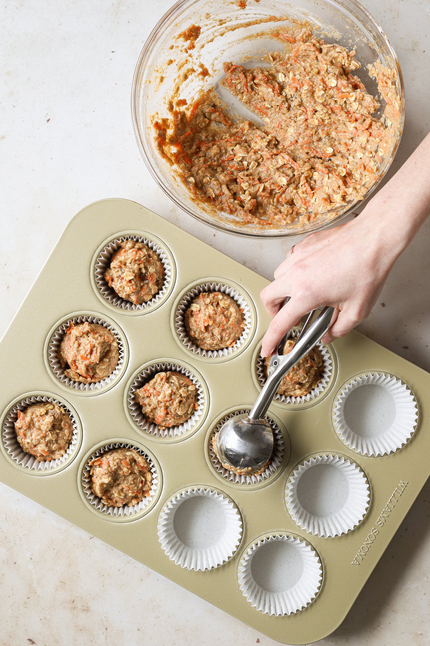 Oatmeal carrot muffin batter being portioned into a lined muffin tin with a small scooper.
