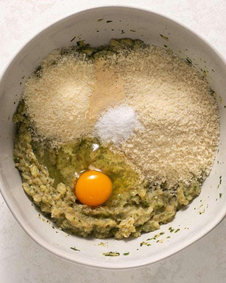 Mashed eggplant in a white bowl with an egg, breadcrumbs, parmesan cheese, and seasoning. 