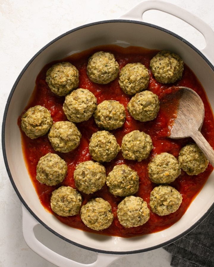 Baked eggplant meatballs in a large white stockpot with pasta sauce and a wooden spoon.