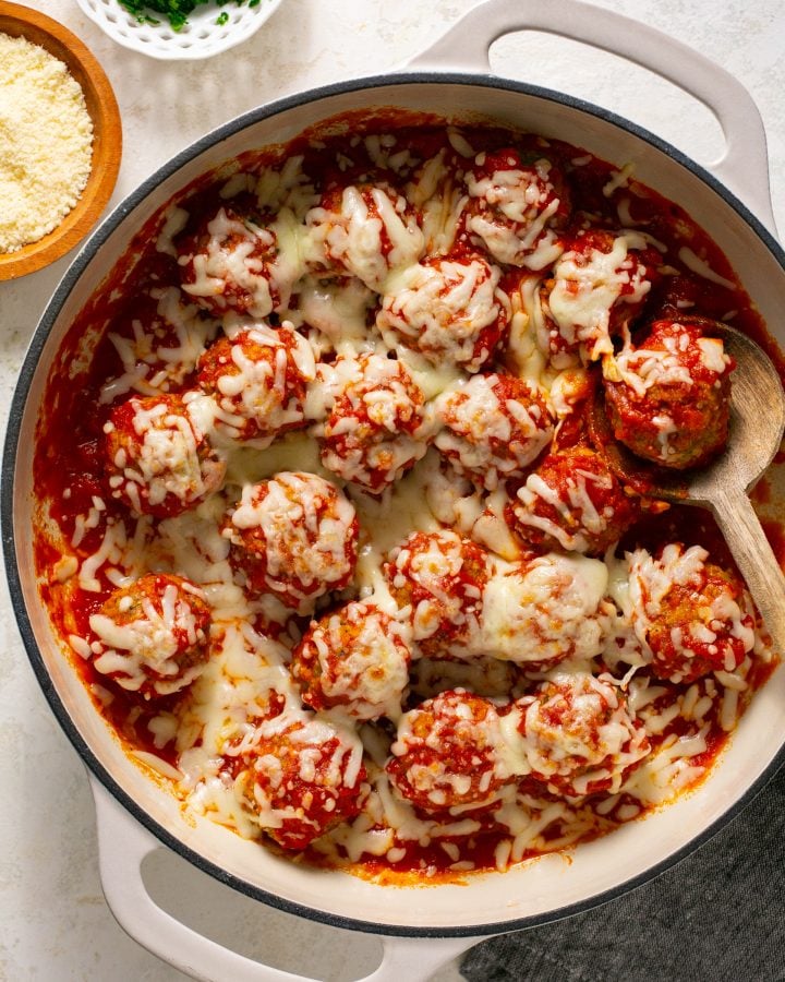 Eggplant meatballs in a stockpot with sauce and a wooden spoon, covered in melted cheese. There is a small bowl of parmesan cheese next to it.