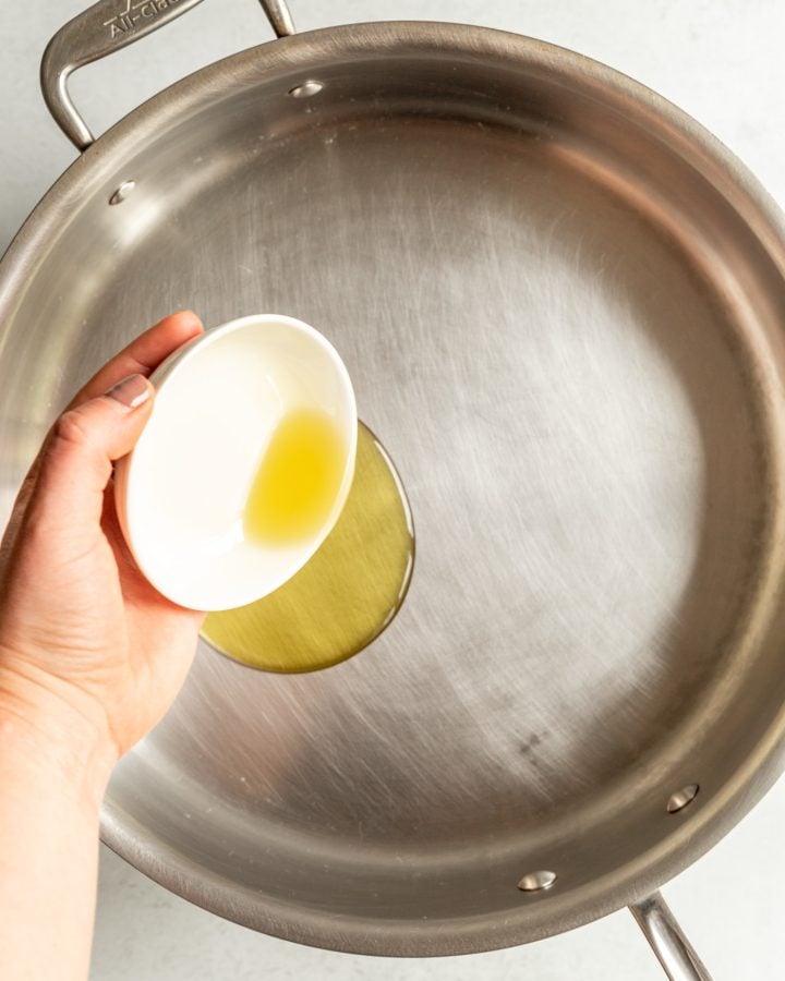 A person pouring oil from a small white bowl into a large skillet.