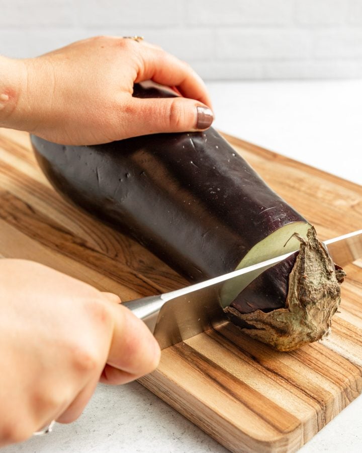A person using a large knife to cut the stem off an eggplant. 