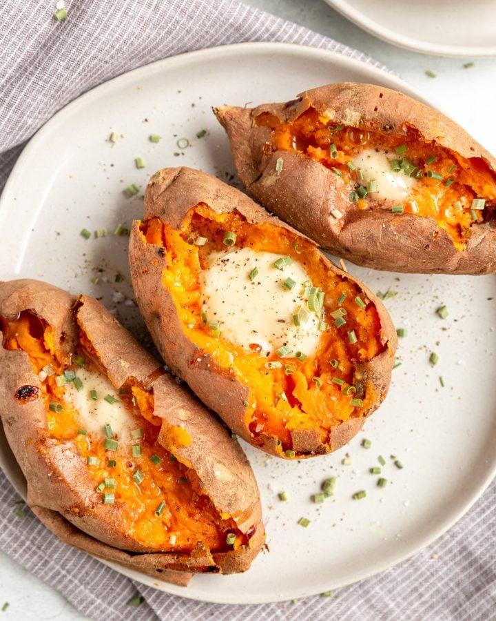 Oven baked sweet potatoes on a round white plate. They are cut open and have melting butter, pepper, and chives sprinkled over them. 