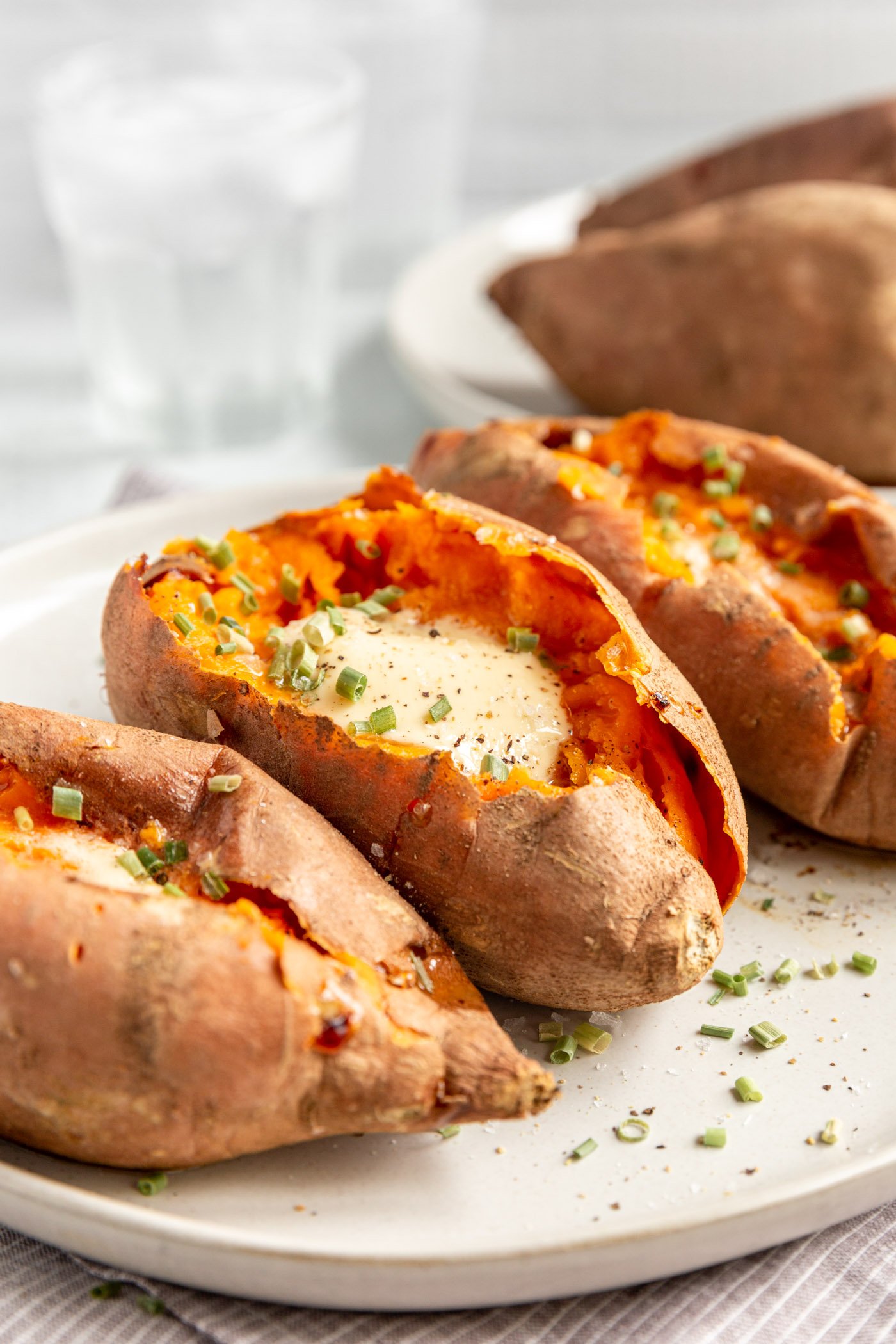 Oven baked sweet potatoes on a plate, cut open and topped with melting butter and chives.