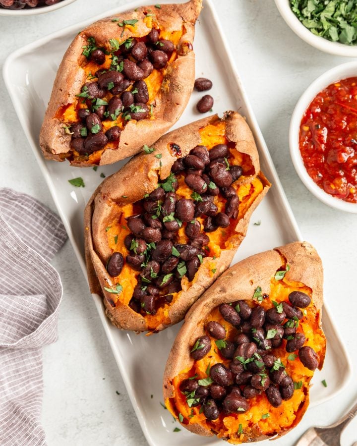 Oven baked sweet potatoes on a white serving plate. They are stuffed with black beans and fresh herbs. There is a napkin and a bowl of salsa next to the platter.