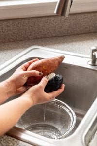 A person washing a sweet potato over the sink with a scrub brush.