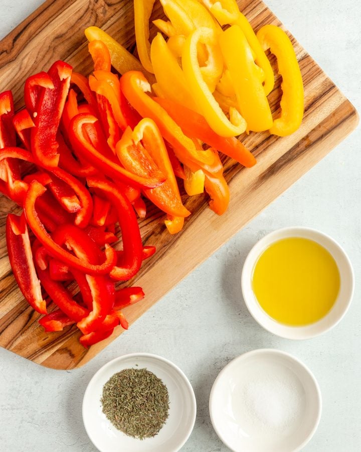 Sliced bell peppers on a wooden cutting board with bowls of dried thyme, salt, and oil next to it. 