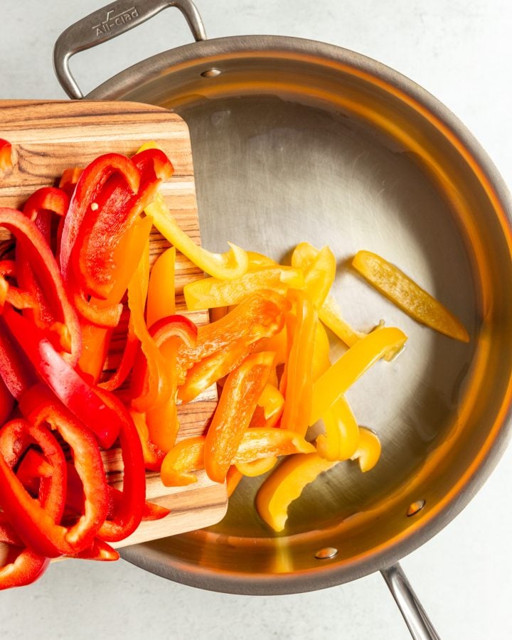 Sliced bell peppers being poured from a wooden cutting board into a large metal skillet with oil.
