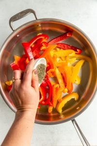 A hand pouring salt and dried thyme into a metal skillet with sliced bell peppers.
