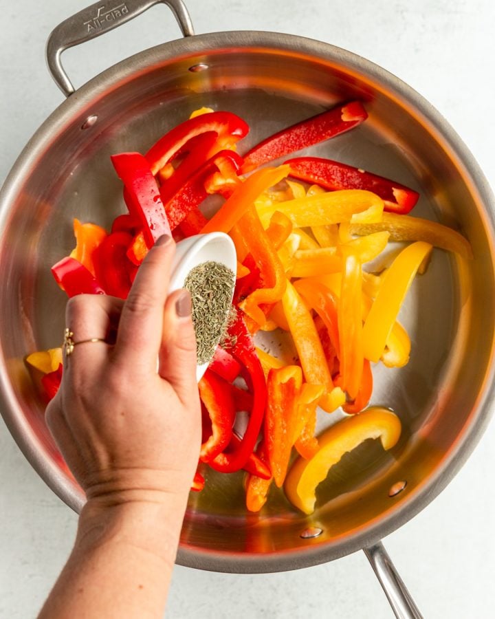 A person pouring dried thyme into a skillet with sliced bell peppers.