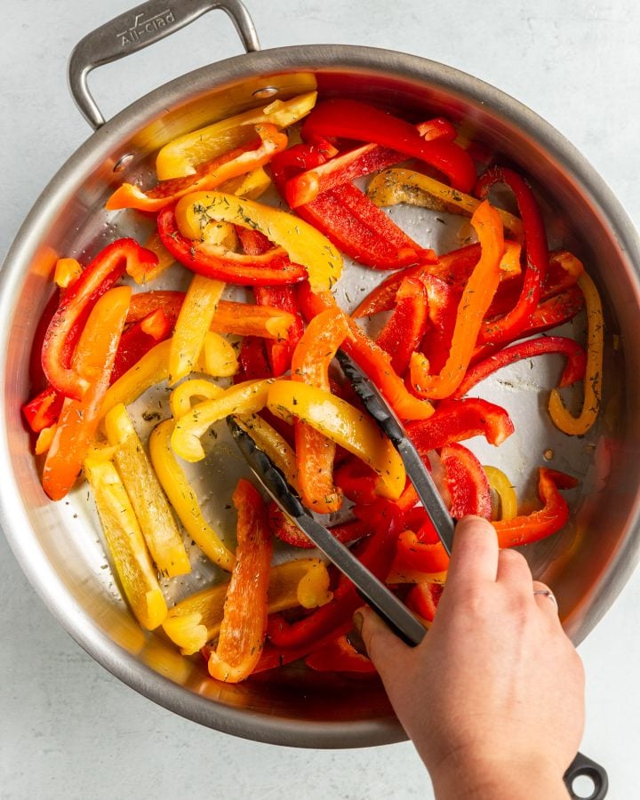 A person using tongs to toss seasoned sliced bell peppers in a skillet. 