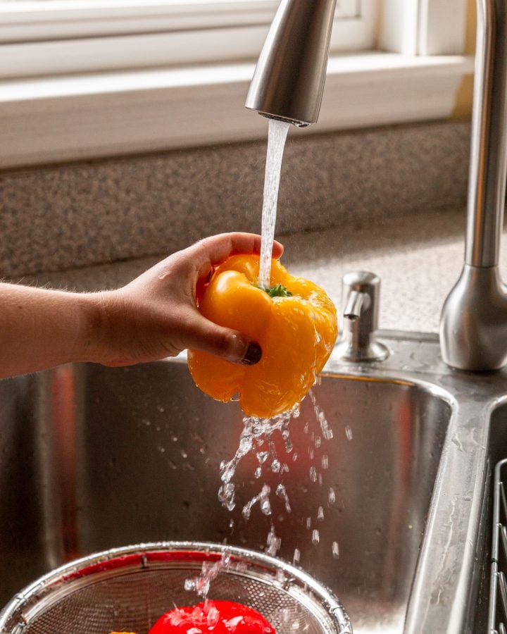 A person washing a bell pepper in a sink over a strainer with more bell peppers.