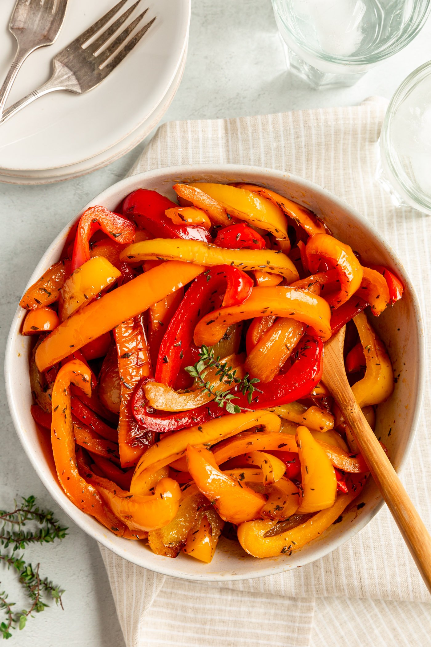Sauteed bell peppers in a bowl with a wooden serving spoon. They are topped with fresh thyme. There is a striped tan napkin under the bowl, fresh thyme next to it, and glassed of water and plates with forks beside it.
