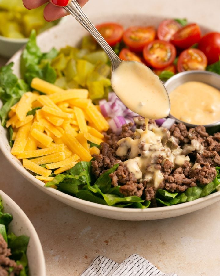 A hand using a spoon to drizzle burger sauce over cheeseburger salad.