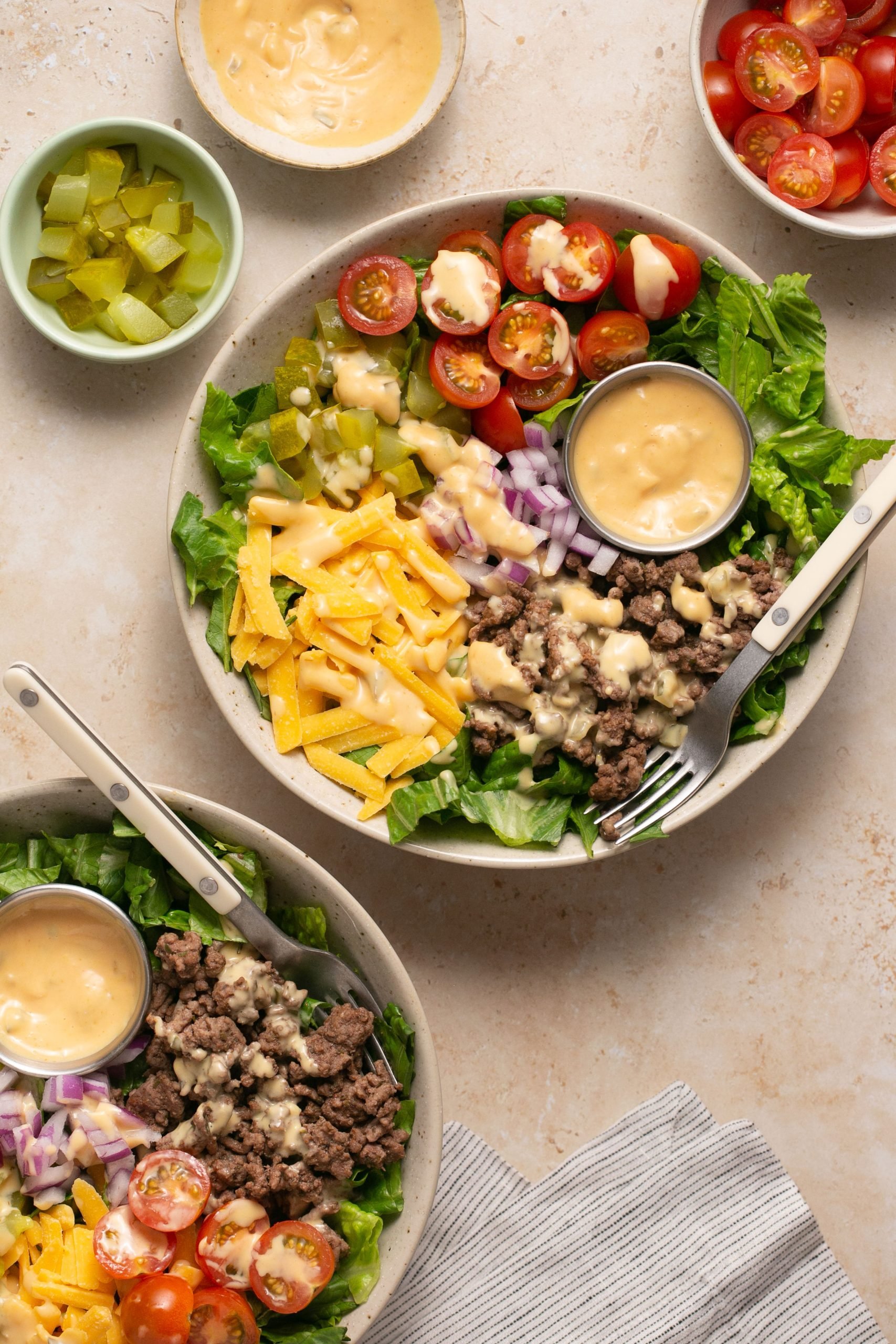 Cheeseburger salads assembled in two bowls on a table with a stripped napkin and two forks. There are small bowls of ingredients on the table too.