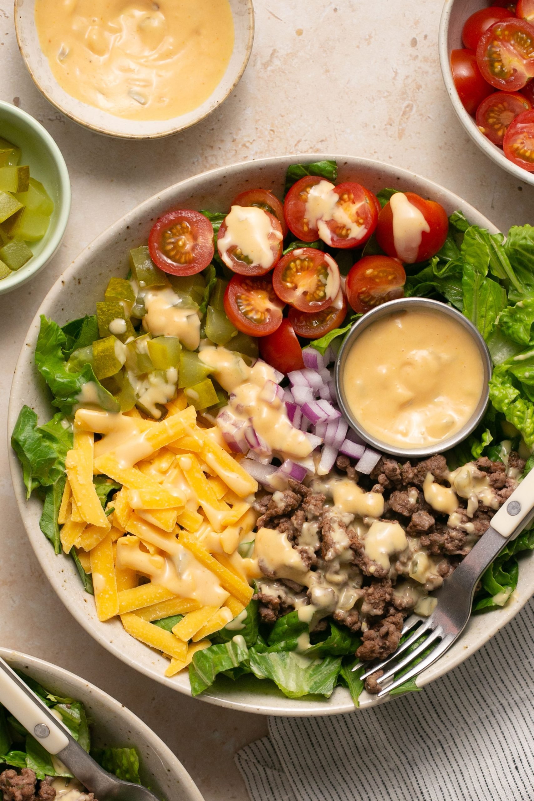 Cheeseburger salad in a bowl with a cup of burger sauce and a fork. Small bowls with ingredients surround the bowl. 