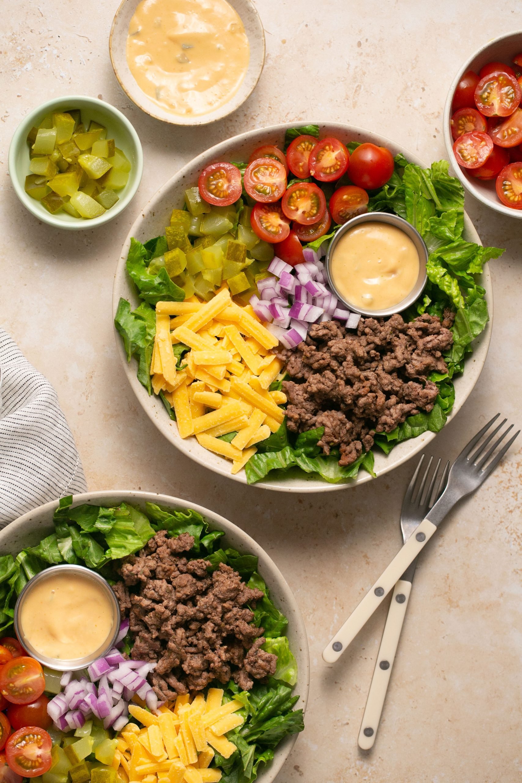 Cheeseburger salads assembled in two bowls on a table with a stripped napkin and two forks. There are small bowls of ingredients on the table too.