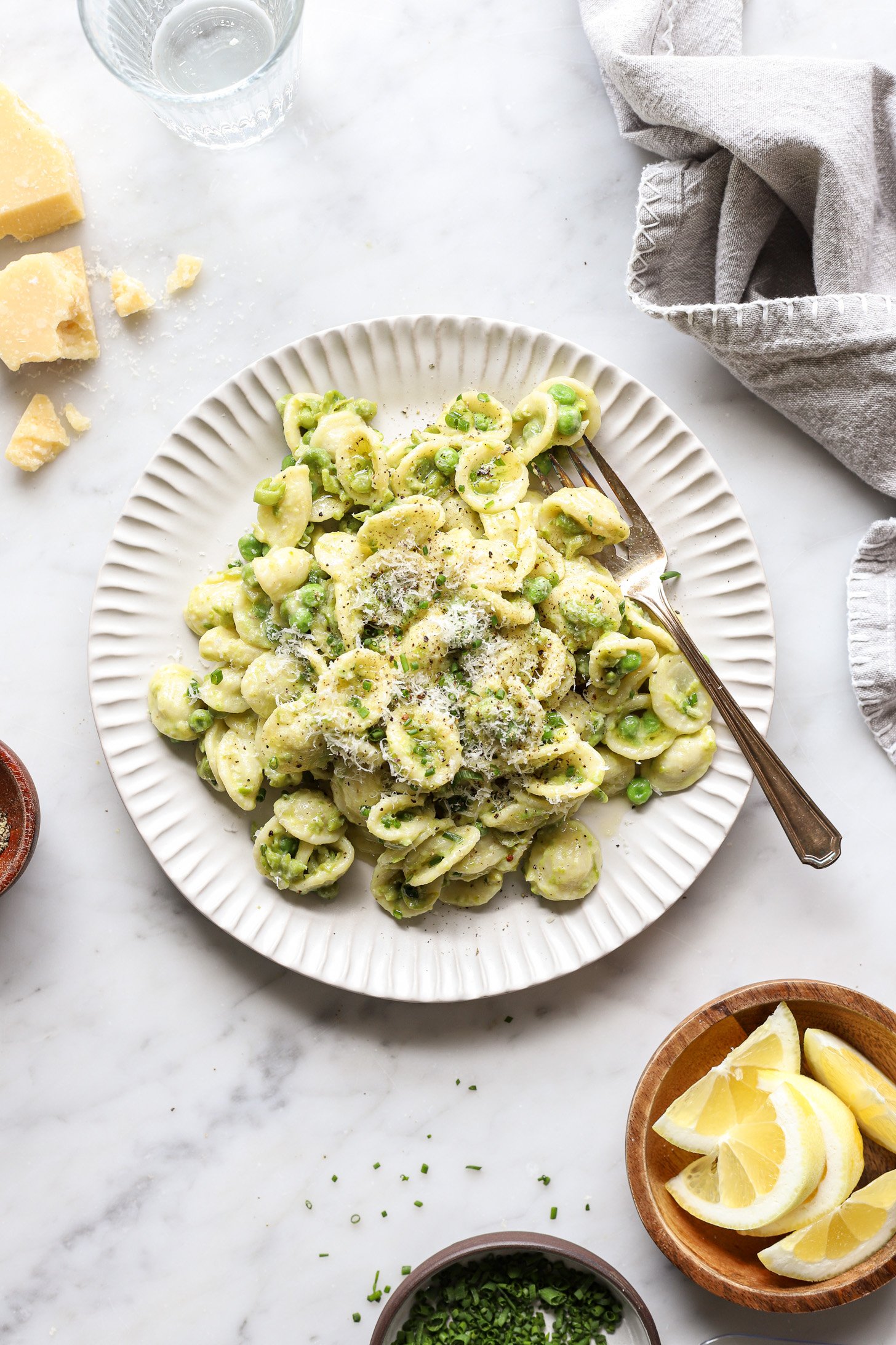 A serving of pasta with peas on a white plate with a fork. The pasta is topped with chives and more parmesan cheese. There is a gray napkin, glass of water, a crumbled block of parmesan cheese , a bowl of sliced lemons and chopped chives and pepper next to the pot on a table. 
