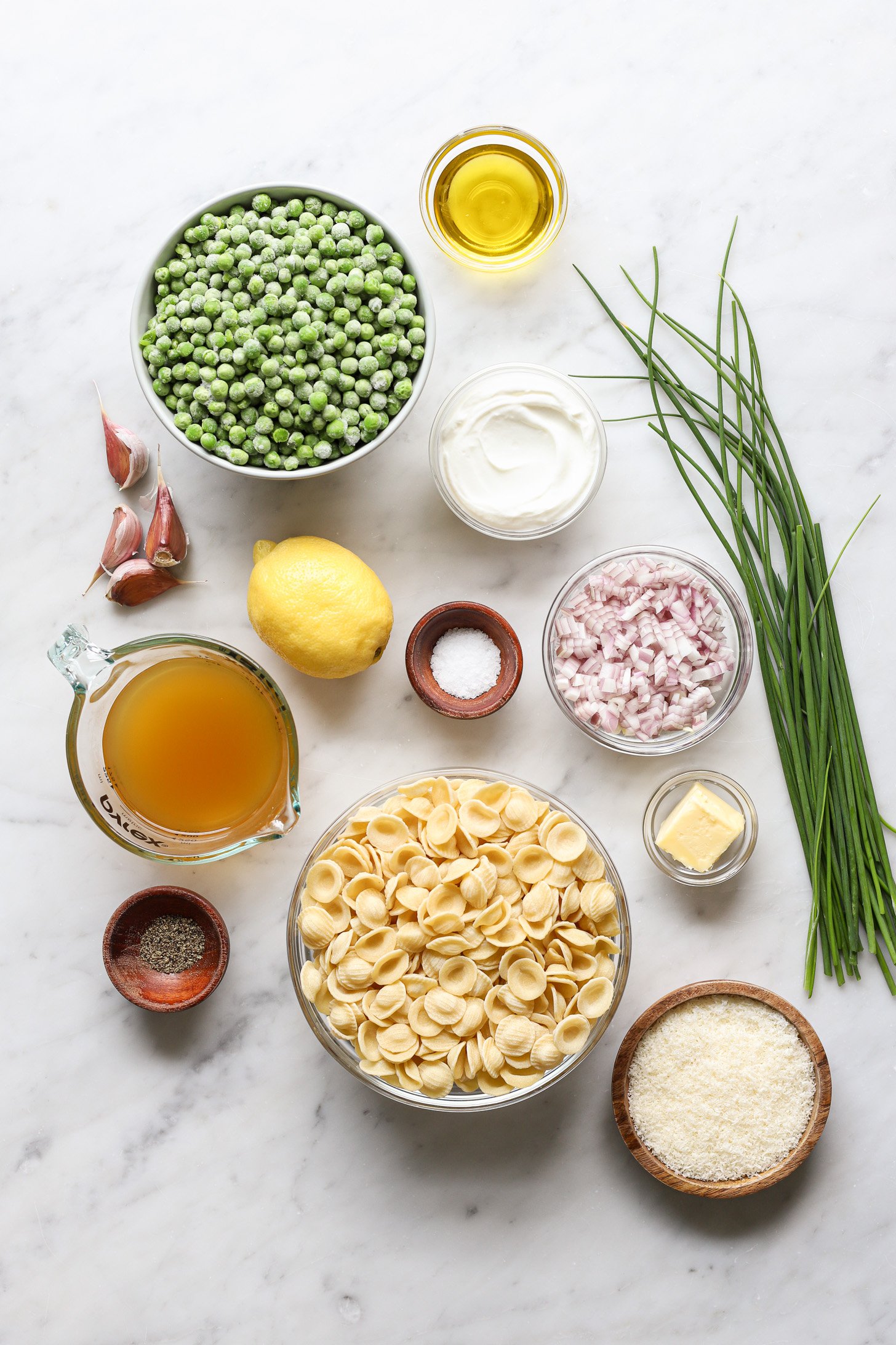 Ingredients for pasta with peas in bowls and measuring cups laid out on a white table. 