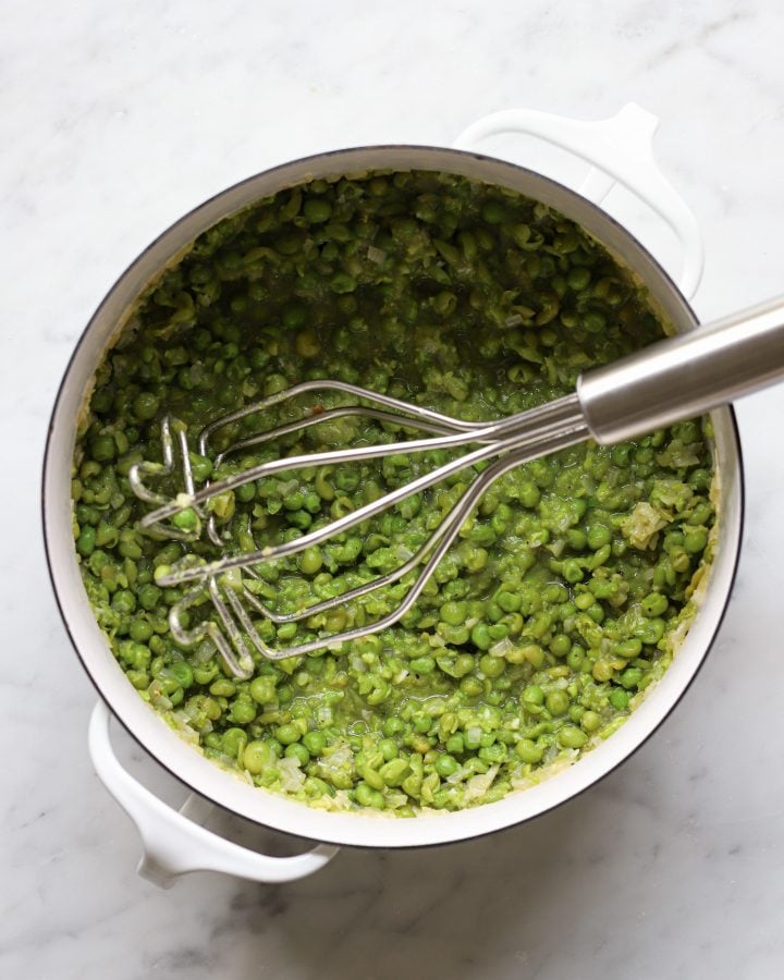 A potato masher in a pot with cooked peas. The peas are mostly mashed.