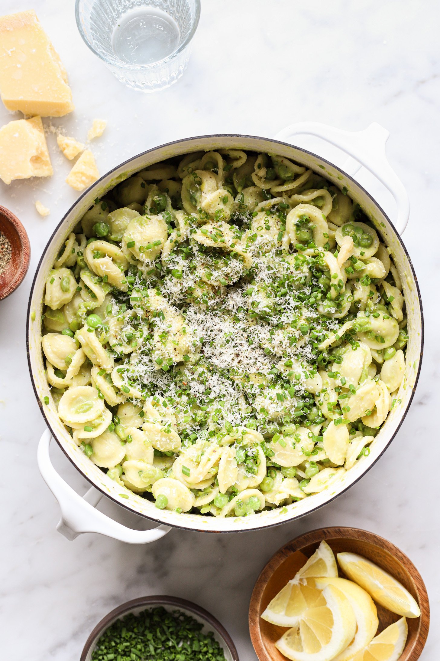 Cooked pasta stirred in to creamy pea sauce in a white pot and topped with chives and more parmesan cheese. There is a glass of water,  a crumbled block of parmesan cheese , a bowl of sliced lemons and chopped chives and pepper next to the pot on a table. 